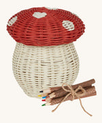 An Olli Ella Porcini mushroom shaped Basket with a red spotted lid, and wooden twig colouring pencils wrapped together with string next to it pictured on a cream background