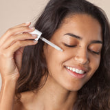 A person applying the Green People Nordic Roots Hyaluronic Booster Serum -onto their face. The image shows the silky, white serum in its lid dropper