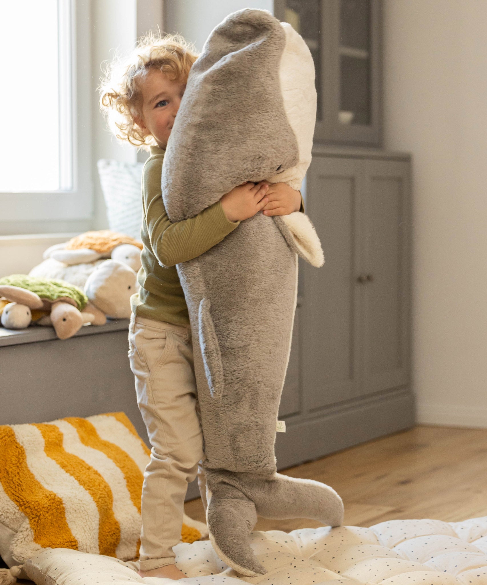 A child playing and holding the Senger Giant grey whale available at Babipur. A soft and cuddly giant whale toy in grey with playful facial features