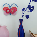 Stripy butterfly wall decoration available at Babipur hanging on a wall above a pink and blue vase. A Studio roof collaboration with artist Zoe Miller.