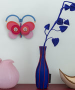 Stripy butterfly wall decoration available at Babipur hanging on a wall above a pink and blue vase. A Studio roof collaboration with artist Zoe Miller.