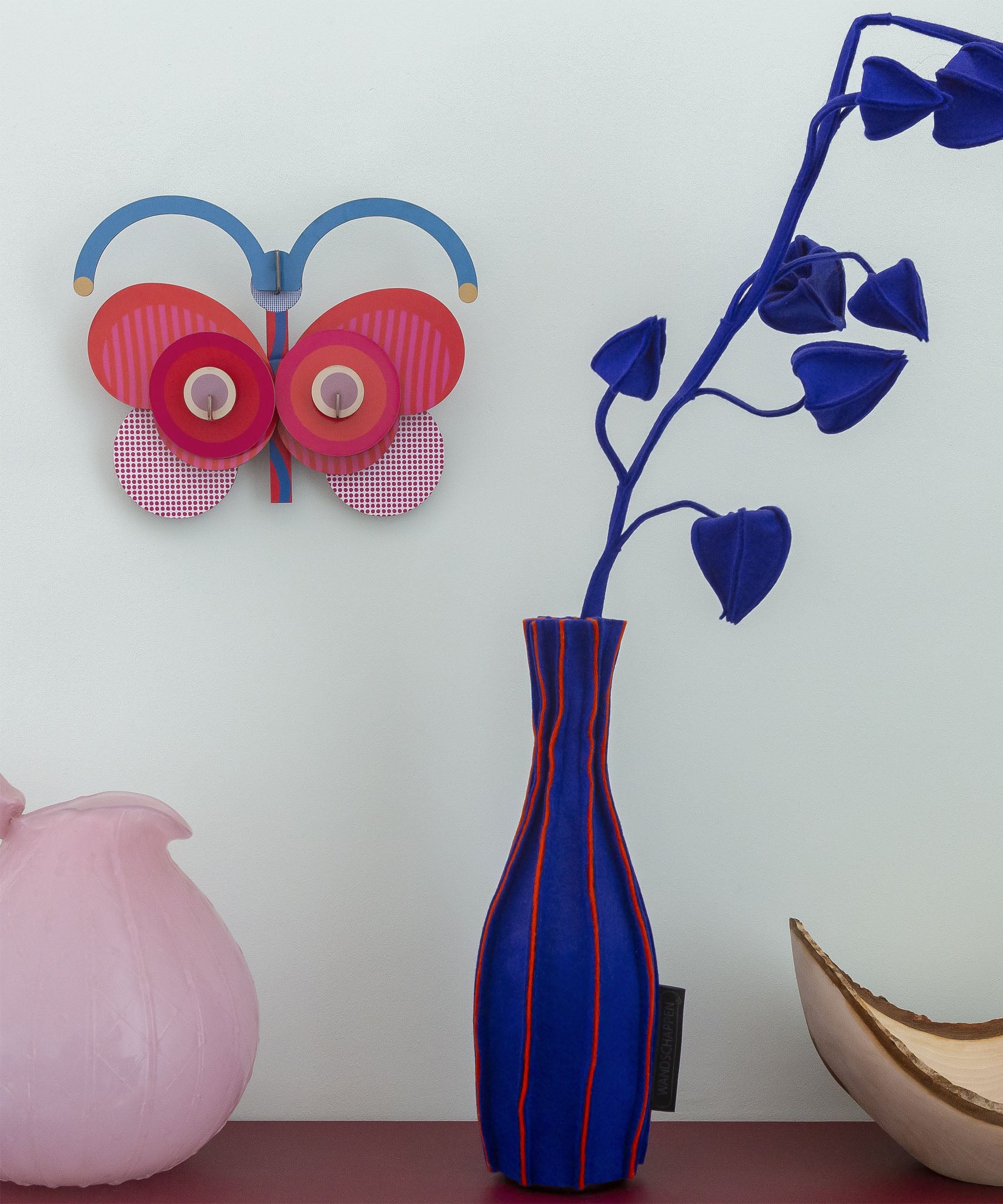 Stripy butterfly wall decoration available at Babipur hanging on a wall above a pink and blue vase. A Studio roof collaboration with artist Zoe Miller.