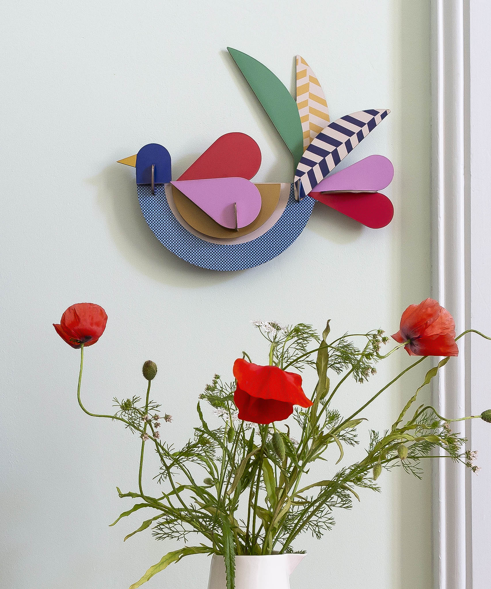 A close up of the feathery bird wall decoration available at Babipur hanging on a wall above red poppies