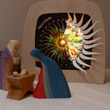 Mary and Joseph with baby Jesus wooden figures next to the Toverlux sonne mond und erde nativity advent calendar available at Babipur. Calendar has windows open revealing a lit up silhouette.