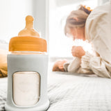 Hevea Glass Baby Bottle Wide Neck With Rubber Sleeve - 150ml in Seafoam Blue on a bed in the foreground with a mother and baby in the background.
