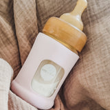 Hevea Glass Baby Bottle Wide Neck With Rubber Sleeve - 150ml in Sand on a taupe muslin blanket.