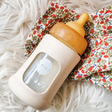 Hevea Glass Baby Bottle Wide Neck With Rubber Sleeve - 150ml in Powder Pink on a white furry rug and floral muslin.