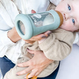Toddler sitting on parents lap drinking from the Hevea Glass Bottle Wide Neck With Rubber Sleeve - 250ml in Seafoam Blue.