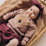 A close up of a baby lying on a fluffy blanket and wearing the Wooly Organic embroidered sweater in pale blush pink /savannah available at Babipur