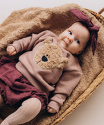 A close up of a baby lying on a fluffy blanket and wearing the Wooly Organic embroidered sweater in pale blush pink /savannah available at Babipur