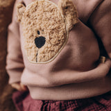 A close up of a baby lying on a fluffy blanket and wearing the Wooly Organic embroidered sweater in pale blush pink /savannah available at Babipur showing the cute fluffy cream teddy bear on the front.