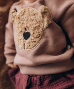 A close up of a baby lying on a fluffy blanket and wearing the Wooly Organic embroidered sweater in pale blush pink /savannah available at Babipur showing the cute fluffy cream teddy bear on the front.