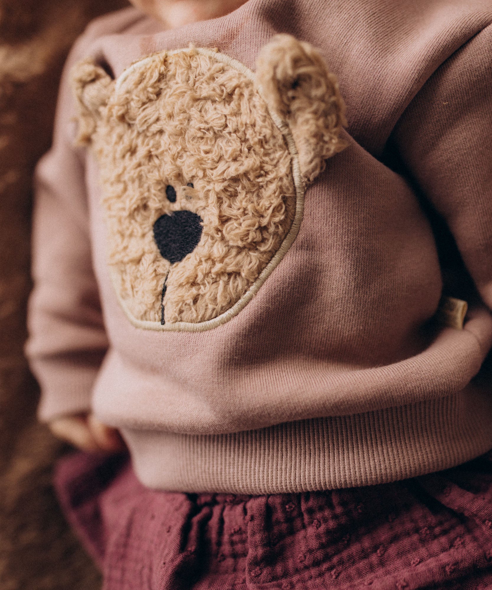 A close up of a baby lying on a fluffy blanket and wearing the Wooly Organic embroidered sweater in pale blush pink /savannah available at Babipur showing the cute fluffy cream teddy bear on the front.