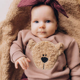 A close up of a baby lying on a fluffy blanket and wearing the Wooly Organic embroidered sweater in pale blush pink /savannah available at Babipur showing the cute fluffy cream teddy bear on the front. Features popper fasteners on the shoulder for a easy fit