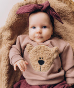 A close up of a baby lying on a fluffy blanket and wearing the Wooly Organic embroidered sweater in pale blush pink /savannah available at Babipur showing the cute fluffy cream teddy bear on the front. Features popper fasteners on the shoulder for a easy fit