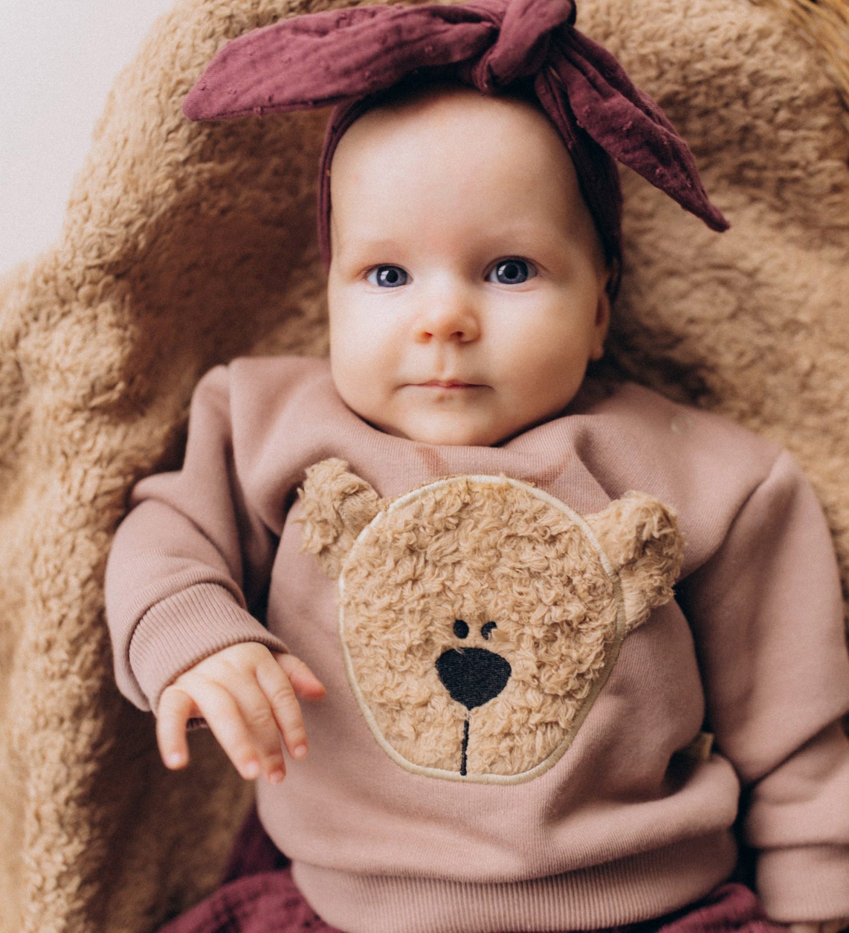 A close up of a baby lying on a fluffy blanket and wearing the Wooly Organic embroidered sweater in pale blush pink /savannah available at Babipur showing the cute fluffy cream teddy bear on the front. Features popper fasteners on the shoulder for a easy fit