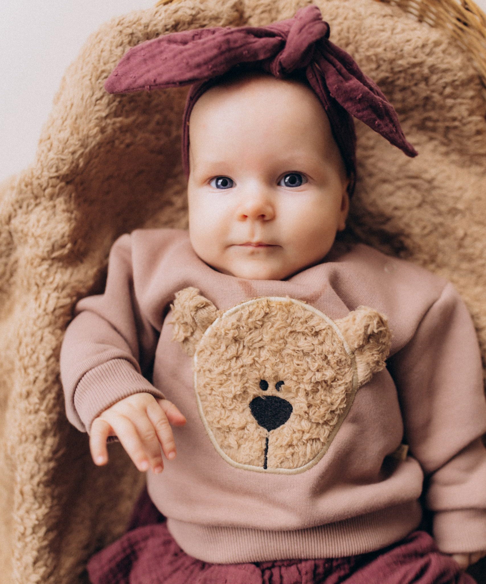 A close up of a baby lying on a fluffy blanket and wearing the Wooly Organic embroidered sweater in pale blush pink /savannah available at Babipur showing the cute fluffy cream teddy bear on the front. Features popper fasteners on the shoulder for a easy fit