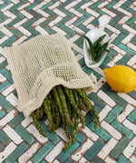 A Slice Of Green small mesh produce bag made from recycled cotton showing a bunch of asparagus inside next a lemon and chillies showing scale.