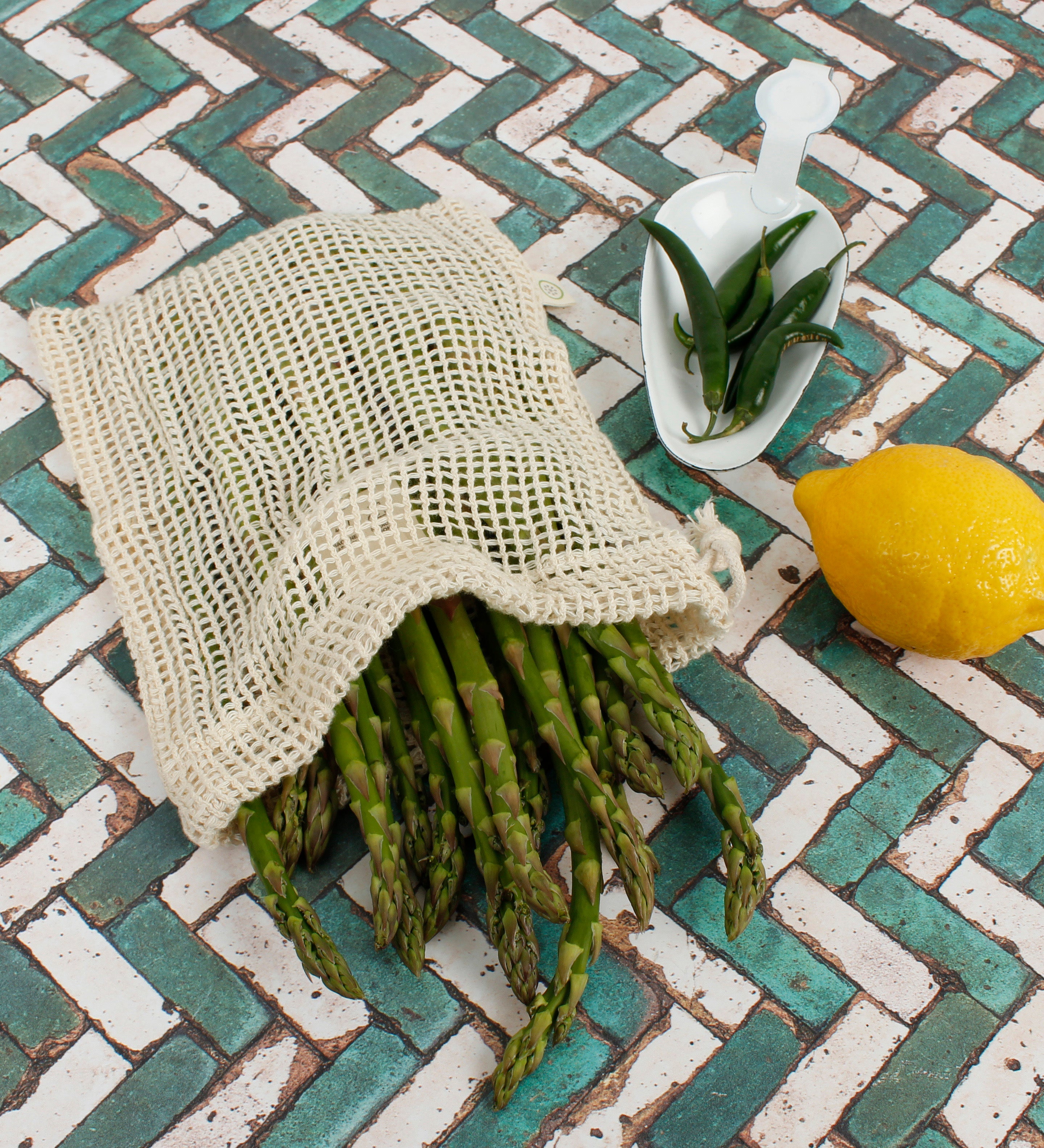 A Slice Of Green small mesh produce bag made from recycled cotton showing a bunch of asparagus inside next a lemon and chillies showing scale.