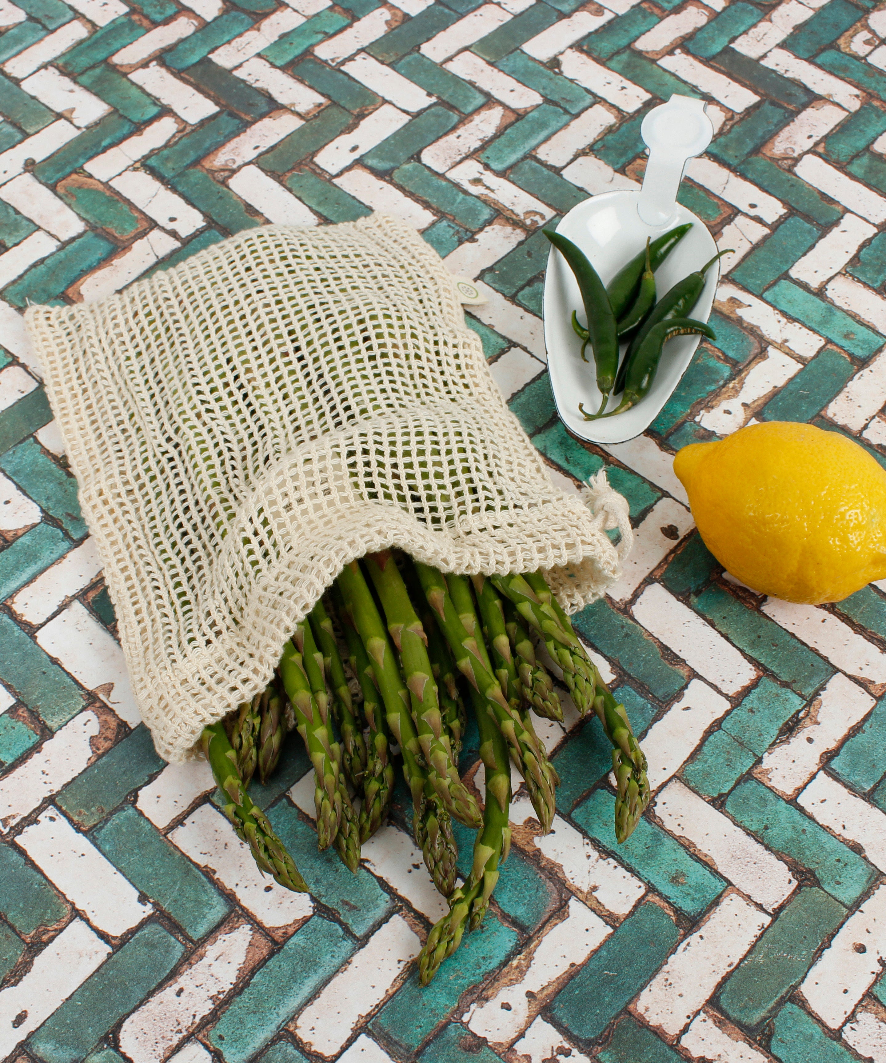 A Slice Of Green small mesh produce bag made from recycled cotton showing a bunch of asparagus inside next a lemon and chillies showing scale.