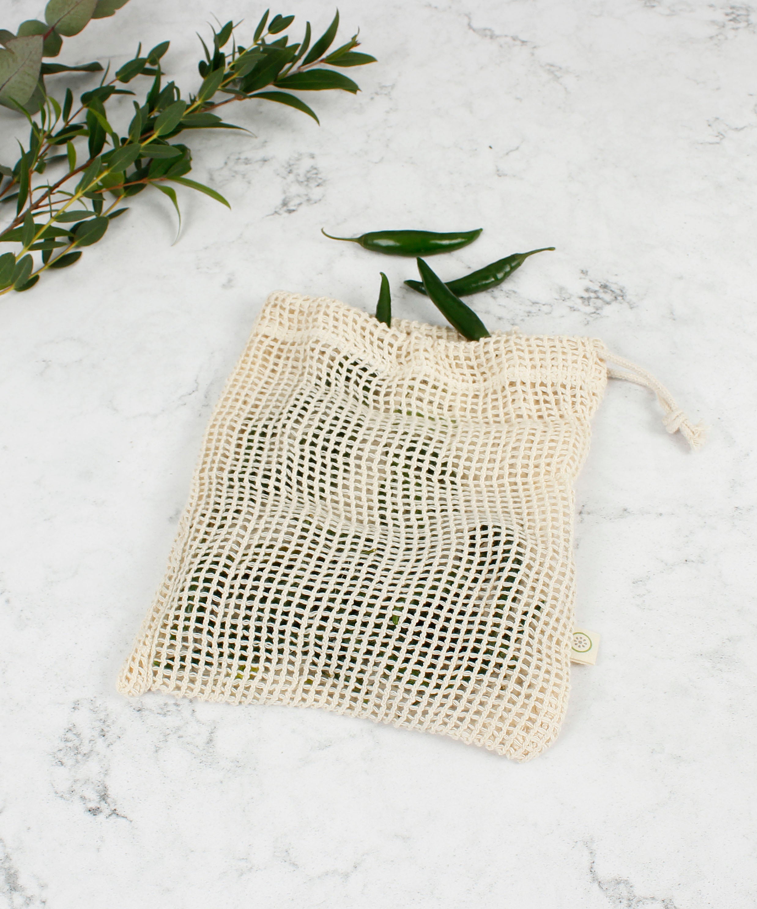 A Slice Of Green small mesh produce bag laid flat on a stone surface. A recycled cotton bag showing a bunch of green chillies inside.