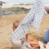 The check blue and white design muslin cloth from the Avery Row Coastline Baby Muslin Squares in a Set being held up by an adults, a baby can be seen sitting behind. The pack has one plain coral coloured cloth, one check blue and white waves design and one coastline print design. Avery Row have a range of stylish baby and nursery essential available here at babipur.