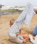 The check blue and white design muslin cloth from the Avery Row Coastline Baby Muslin Squares in a Set being held up by an adults, a baby can be seen sitting behind. The pack has one plain coral coloured cloth, one check blue and white waves design and one coastline print design. Avery Row have a range of stylish baby and nursery essential available here at babipur.