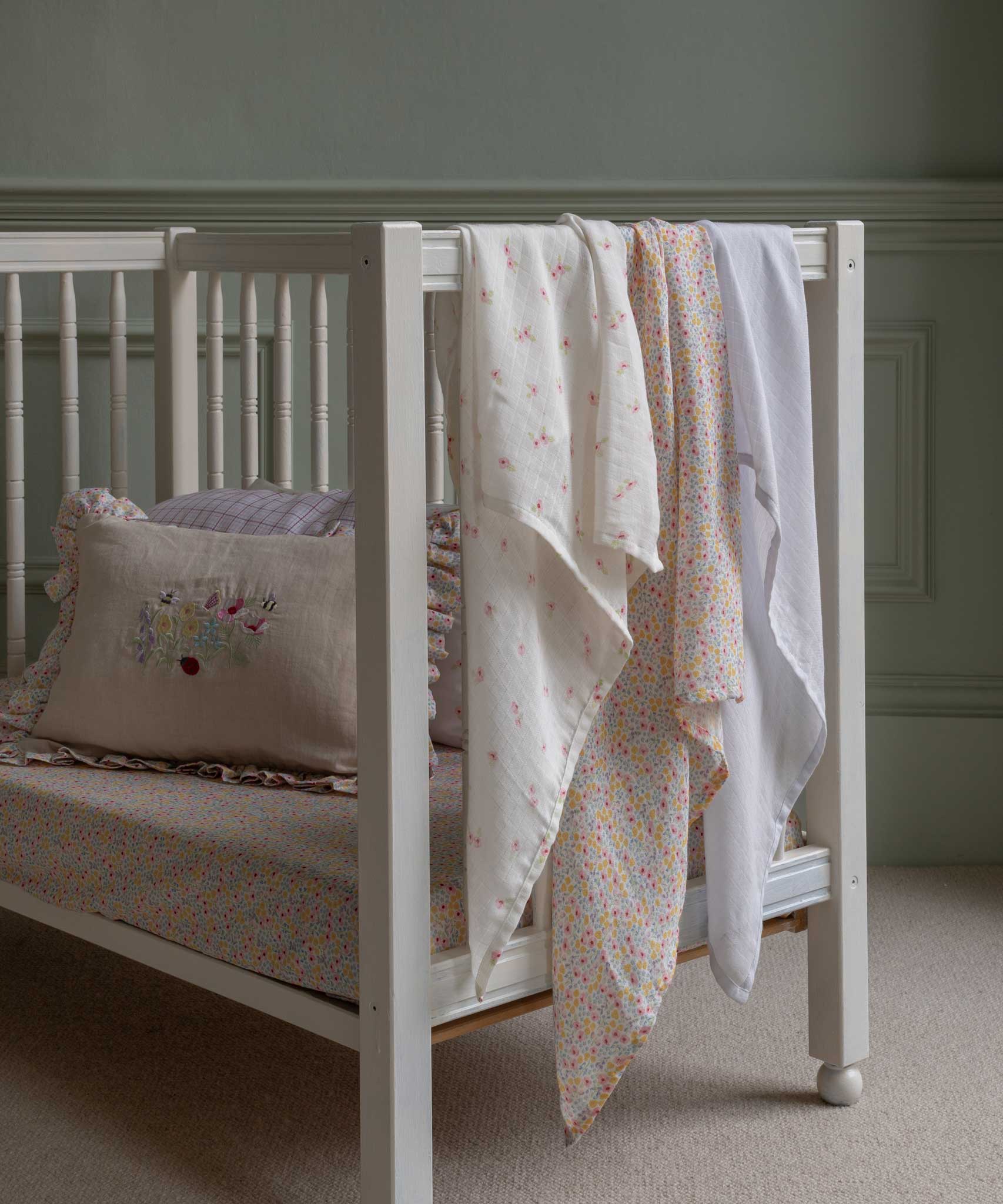 The cloths from the Avery Row Wildflower Baby Muslin Squares in a Set of 3 draped on the side of a cot bed. One cloth is plain white, one has delicate pink flowers and one has an all over floral wildflower design. Avery Row have a collection of stylish nursery and baby essentials available at Babipur.
