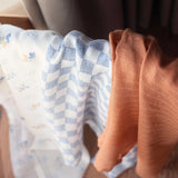 Avery Row Coastline Baby Muslin Squares in a Set of 3. The cloths have been placed on stand. The pack has one plain coral coloured cloth, one check blue and white waves design and one coastline print design. Avery Row have a range of stylish baby and nursery essential available here at babipur.