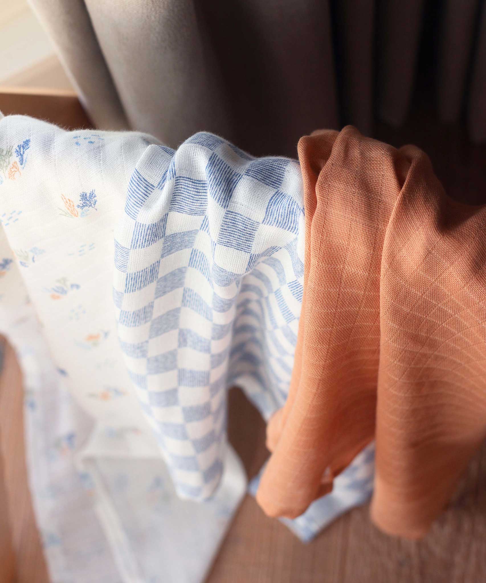 Avery Row Coastline Baby Muslin Squares in a Set of 3. The cloths have been placed on stand. The pack has one plain coral coloured cloth, one check blue and white waves design and one coastline print design. Avery Row have a range of stylish baby and nursery essential available here at babipur.