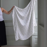 Avery Row Coastline baby Muslin Swaddle. An adult can be seen standing to the side holding up the large swaddle. The swaddle features a delicate coastline print that has corals and little blue fish. Avery Row have a collection of stylish nursery and baby essentials available at Babipur.