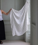 Avery Row Coastline baby Muslin Swaddle. An adult can be seen standing to the side holding up the large swaddle. The swaddle features a delicate coastline print that has corals and little blue fish. Avery Row have a collection of stylish nursery and baby essentials available at Babipur.