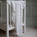 Avery Row Coastline baby Muslin Swaddle. The muslin has been draped over the side of a white cot bed. The swaddle features a delicate coastline print that has corals and little blue fish. Avery Row have a collection of stylish nursery and baby essentials available at Babipur.