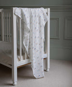 Avery Row Coastline baby Muslin Swaddle. The muslin has been draped over the side of a white cot bed. The swaddle features a delicate coastline print that has corals and little blue fish. Avery Row have a collection of stylish nursery and baby essentials available at Babipur.