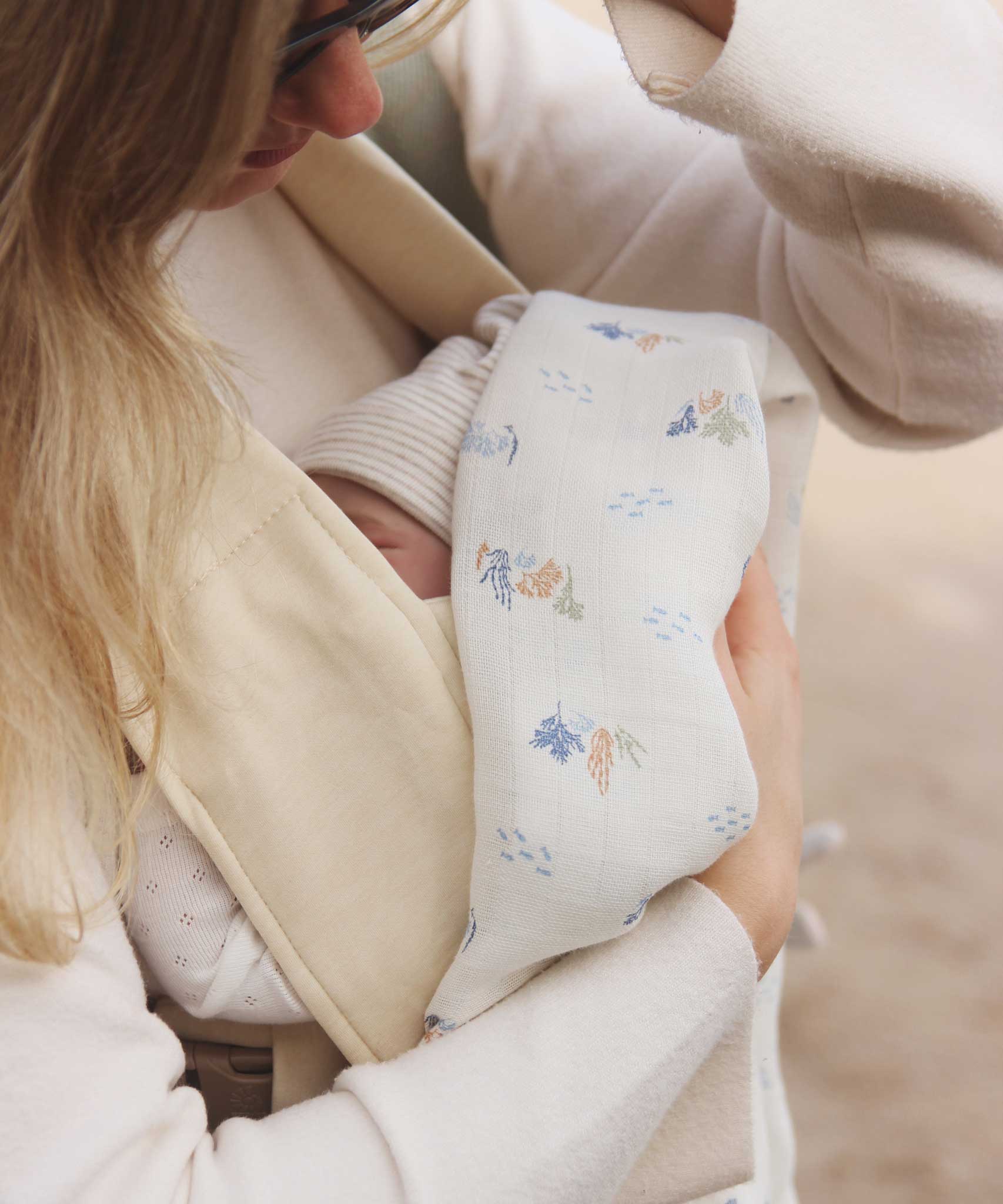 A close up of a baby in a baby carrier, a woman can be seen holding a swaddle that is covering the baby  Avery Row Coastline baby Muslin Swaddle. The swaddle features a delicate coastline print that has corals and little blue fish. Avery Row have a collection of stylish nursery and baby essentials available at Babipur.