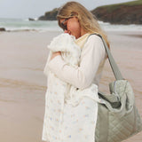 Avery Row Coastline baby Muslin Swaddle. A woman can be seen baby wearing and holding the muslin swaddle over the baby in the carrier. The swaddle features a delicate coastline print that has corals and little blue fish. Avery Row have a collection of stylish nursery and baby essentials available at Babipur.
