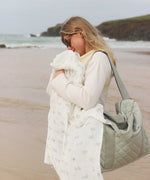 Avery Row Coastline baby Muslin Swaddle. A woman can be seen baby wearing and holding the muslin swaddle over the baby in the carrier. The swaddle features a delicate coastline print that has corals and little blue fish. Avery Row have a collection of stylish nursery and baby essentials available at Babipur.