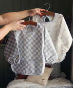 An adults hand holding a hanger that has an Avery Row Sleeved Bib with a check Waves design hung on it. Another design can be seen in the background. This blue and white coloured bib has long sleeves with elasticated cuffs and features a pocket to help catch any food debris. We have a wide range of Avery Row stylish baby products available at Babipur.