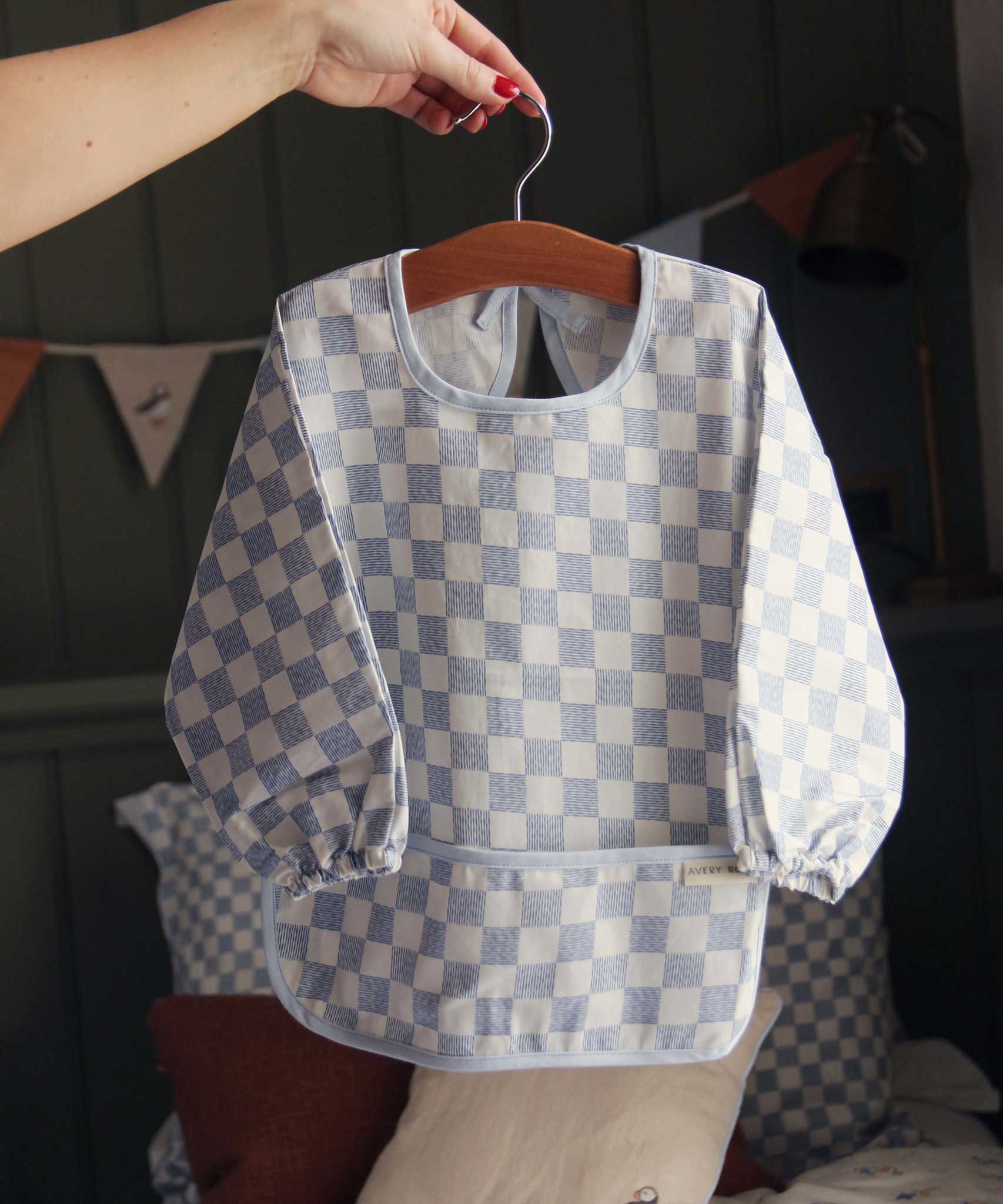 An adults hand holding a hanger that has an Avery Row Sleeved Bib with a check Waves design hung on it. This blue and white coloured bib has long sleeves with elasticated cuffs and features a pocket to help catch any food debris. We have a wide range of Avery Row stylish baby products available at Babipur.