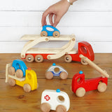 Hand reaching down to play with the Bajo handmade wooden transporter lorry and emergency vehicle toys on a wooden table