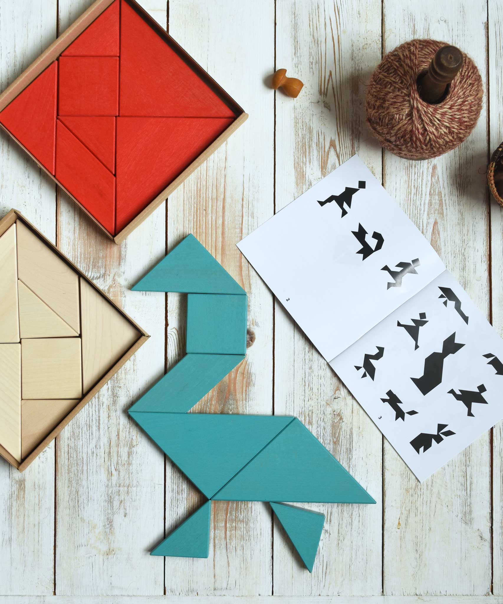 A blue Bajo Tangram puzzle placed on a white wooden surface. The puzzle has been placed in the shape of a bird. The accompanying ideas booklet can be seen open besides it with the red and natural tangram puzzle placed the other side. These heirloom quality toys by Bajo are part of a wide range of wooden toys available here at Babipur.