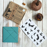 The blue Bajo Tangram wooden toy puzzle placed on a white wooden surface. The pieces of the puzzle have been placed in a square shape next to the packaging and accompanying ideas booklet. These heirloom quality toys by Bajo are part of a wide range of wooden toys available here at Babipur.