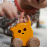 A Bajo push along eye ghost wooden toy on carpet, a child can be seen in the background. The toy has a yellow painted body and natural wooden wheels. These heirloom quality toys by Bajo are part of a wide range of wooden toys available here at Babipur.