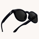 Bird Eyewear Kid's Birdies Sunglasses - Matte Black. A matte black pair of kids sunglasses with a small white bird silhouette on the arm, and black lenses on a cream background