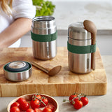 Two Black + Blum insulated stainless steel food flasks placed on a bamboo chopping board. The lid of one flask is open. There are cherry tomatoes in front of them.