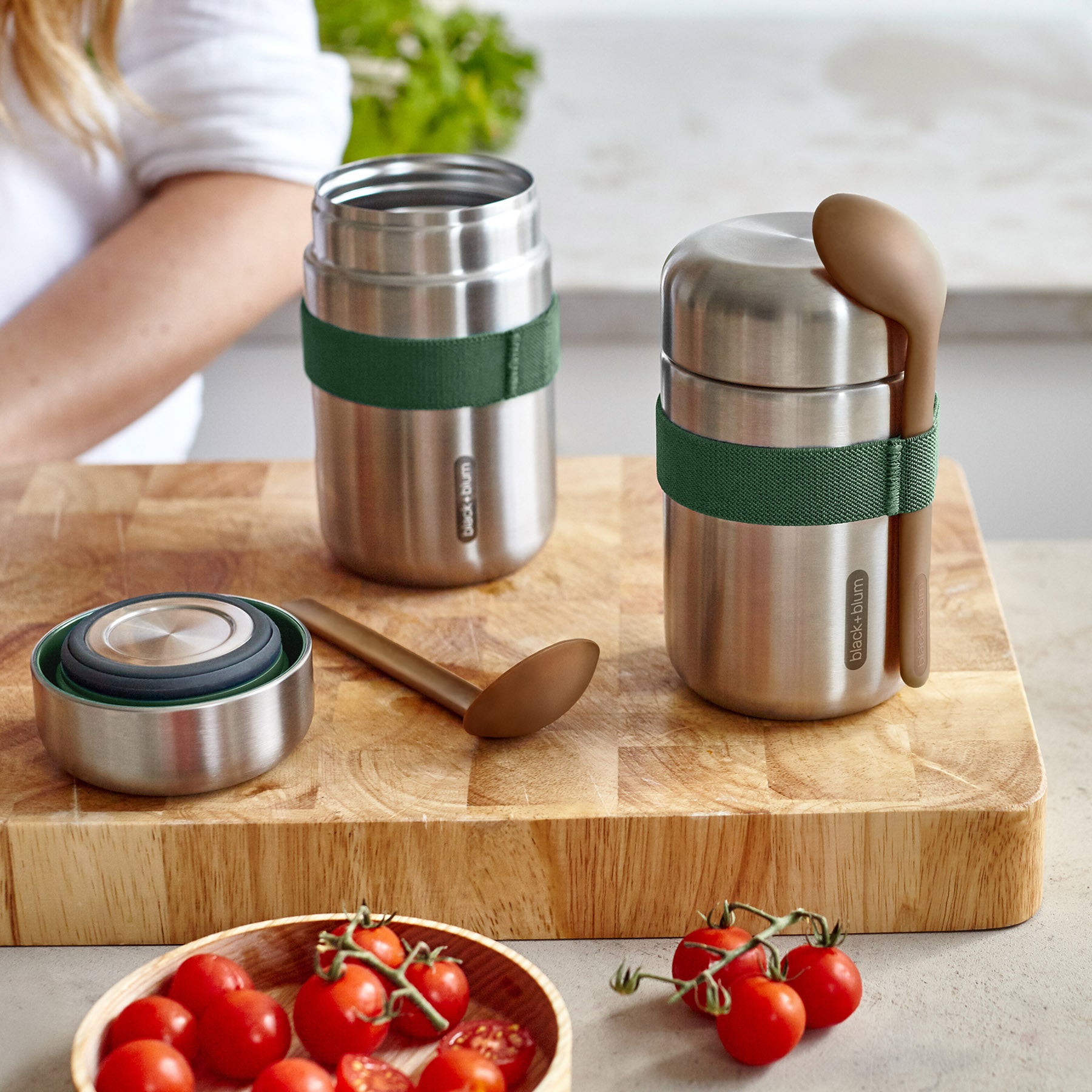 Two Black + Blum insulated stainless steel food flasks placed on a bamboo chopping board. The lid of one flask is open. There are cherry tomatoes in front of them.