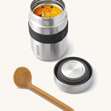 A close up of the Black + Blum insulated stainless steel food flask with wooden spoon showing the black elastic strap and lid detail