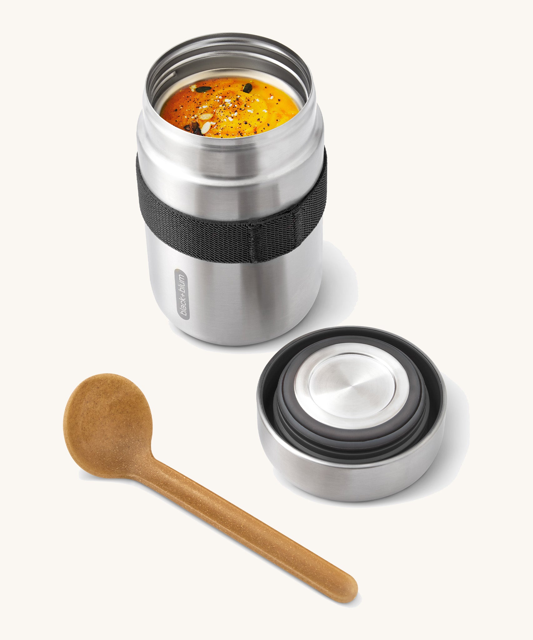 A close up of the Black + Blum insulated stainless steel food flask with wooden spoon showing the black elastic strap and lid detail