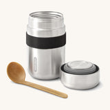 A close up of the Black + Blum insulated stainless steel food flask in black showing the wooden spoon and lid resting next to the flask