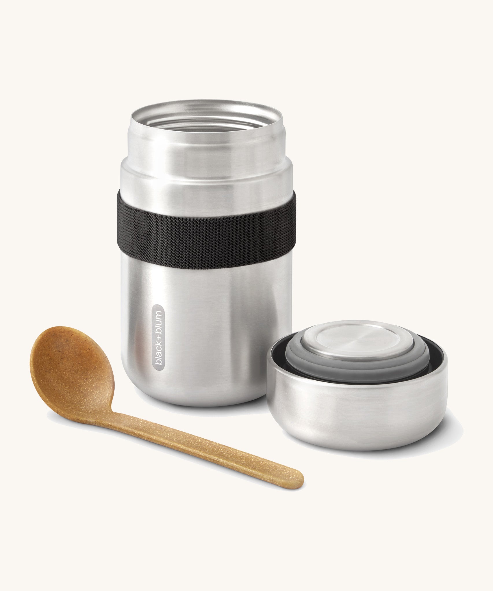A close up of the Black + Blum insulated stainless steel food flask in black showing the wooden spoon and lid resting next to the flask